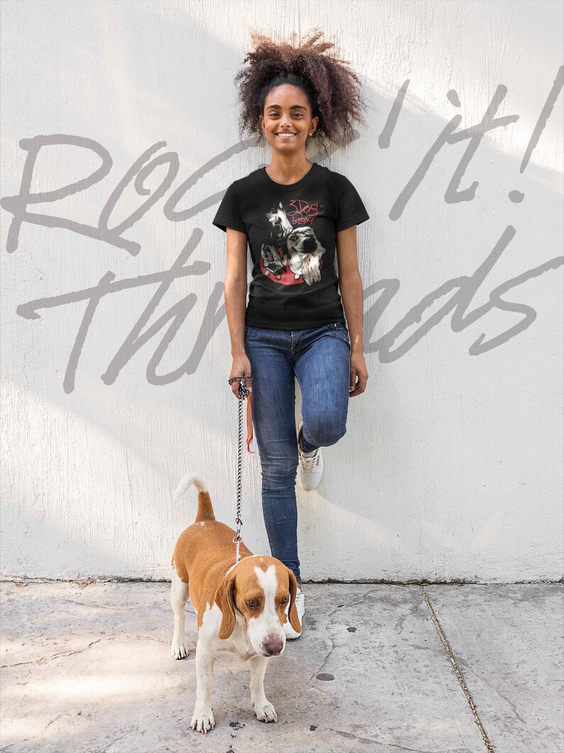 May include: A woman wearing a black t-shirt with a graphic of three dogs and the text "3 Dogs Night" is walking a brown and white beagle on a leash. The woman is standing in front of a white wall with the text "Rock It! Threads" written on it.