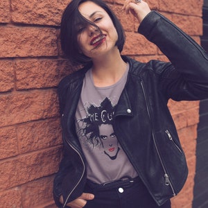 May include: A woman wearing a black leather jacket and a grey t-shirt with a black and white graphic print of a person with the text "The Cure" on it.