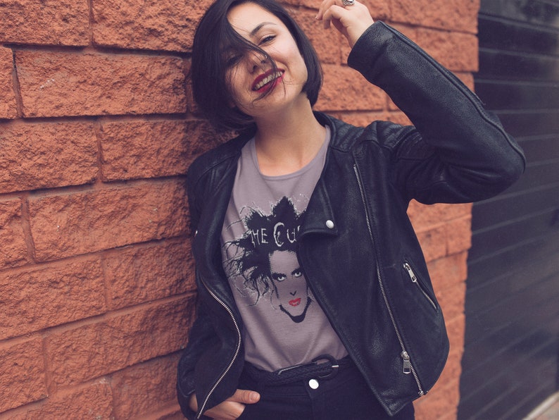May include: A woman wearing a black leather jacket and a grey t-shirt with a black and white graphic print of a person with the text "The Cure" on it.