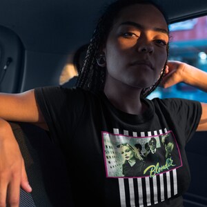 Op de afbeelding: Een zwart T-shirt met een grafische print van de band Blondie. De print heeft een zwart-wit gestreepte achtergrond met een roze rand. De bandleden zijn afgebeeld in de print, met de bandnaam in neon groene letters.