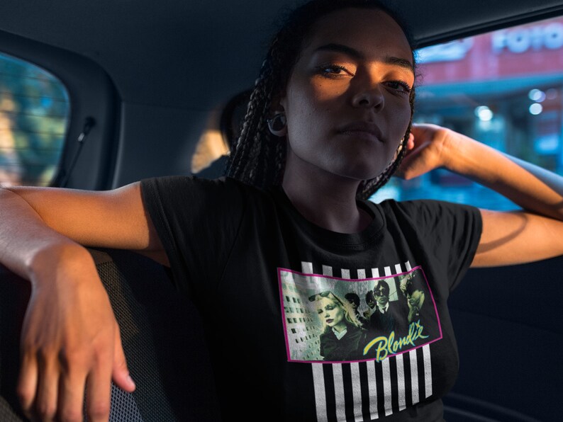 Op de afbeelding: Een zwart T-shirt met een grafische print van de band Blondie. De print heeft een zwart-wit gestreepte achtergrond met een roze rand. De bandleden zijn afgebeeld in de print, met de bandnaam in neon groene letters.