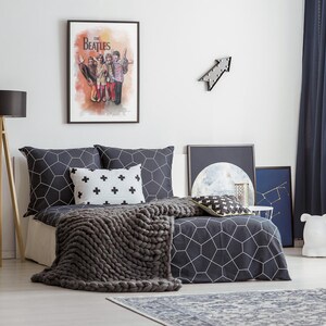 K&ouml;nnte beinhalten: Ein Schlafzimmer mit einem Bett, das mit dunkelgrauer Bettw&auml;sche mit wei&szlig;em geometrischem Muster, einem wei&szlig;en Kissen mit schwarzen Pluszeichen und einer grob gestrickten grauen Decke ausgestattet ist. Ein gerahmtes Beatles-Poster h&auml;ngt an der Wand.