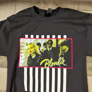 Op de afbeelding: Zwart T-shirt met een wit en zwart gestreept patroon. Het shirt heeft een afbeelding van de band Blondie voor een stadsbeeld. De bandleden dragen zwarte kleding en zonnebrillen. De naam van de band is geschreven in gele en roze letters.