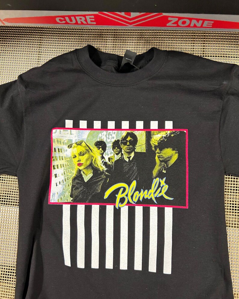 Op de afbeelding: Zwart T-shirt met een wit en zwart gestreept patroon. Het shirt heeft een afbeelding van de band Blondie voor een stadsbeeld. De bandleden dragen zwarte kleding en zonnebrillen. De naam van de band is geschreven in gele en roze letters.
