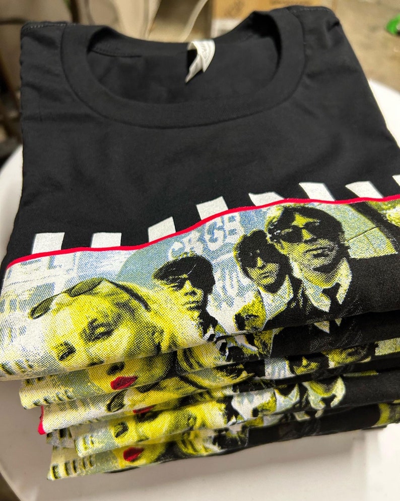 Op de afbeelding: Een stapel zwarte T-shirts met een grafische print van een band. De afbeelding toont een zwart-witte afbeelding van een band tegen een gele en witte achtergrond. De naam van de band is in witte letters gedrukt op een rood-wit gestreepte band.