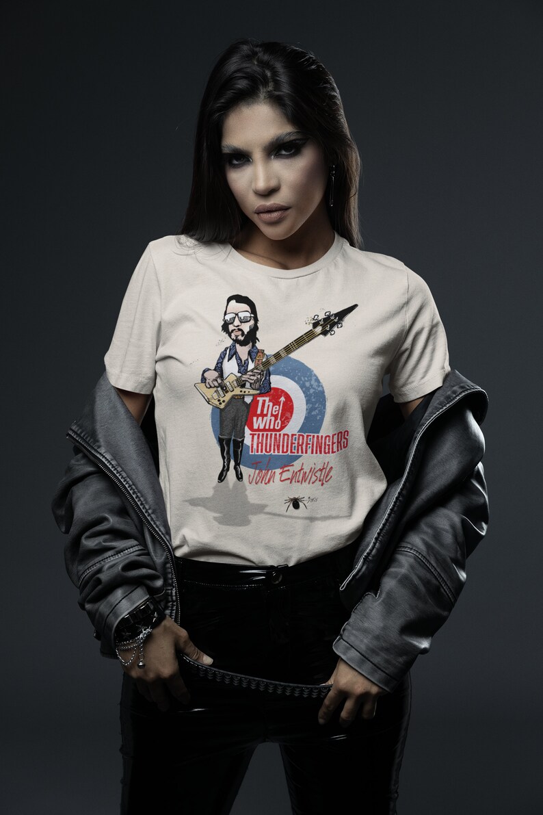 May include: A woman wearing a white t-shirt with a graphic of John Entwistle from The Who, playing a guitar. The graphic includes the text "The Who Thunderfingers John Entwistle".