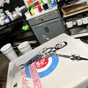 May include: A white t-shirt with a black and white illustration of a person playing a guitar. The text "The Who's Thunderfingers" is printed on the shirt. The shirt is on a table with paint buckets and other supplies.