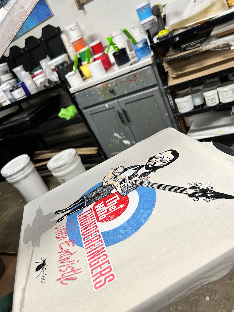 May include: A white t-shirt with a black and white illustration of a person playing a guitar. The text "The Who's Thunderfingers" is printed on the shirt. The shirt is on a table with paint buckets and other supplies.