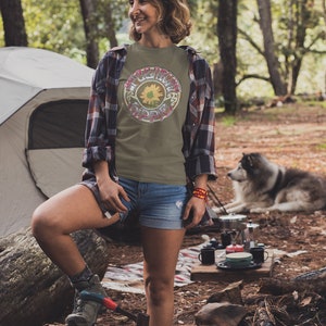 May include: A woman wearing a green t-shirt with a sunflower and the text "The Grateful Dead" in a circle. She is standing in a forest with a tent and a dog in the background.