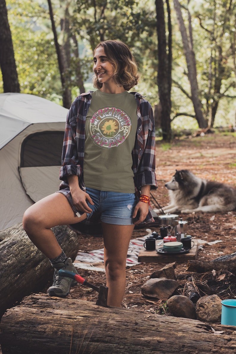 May include: A woman wearing a green t-shirt with a sunflower and the text "The Grateful Dead" in a circle. She is standing in a forest with a tent and a dog in the background.