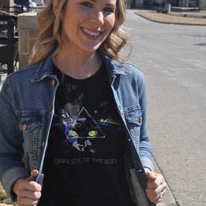 May include: A woman wearing a black t-shirt with a graphic of a cat in a triangle and the text "Dark Side of the Sofa". She is wearing a blue denim jacket over the t-shirt.