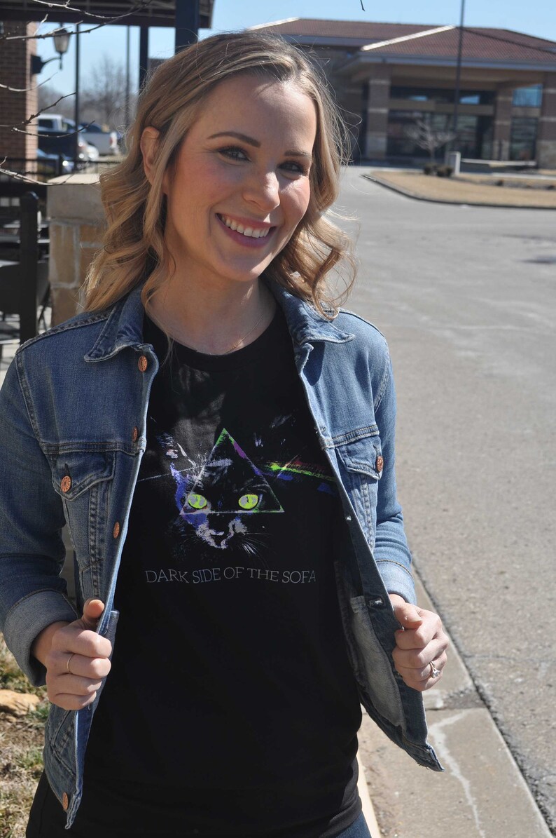 May include: A woman wearing a black t-shirt with a graphic of a cat in a triangle and the text "Dark Side of the Sofa". She is wearing a blue denim jacket over the t-shirt.