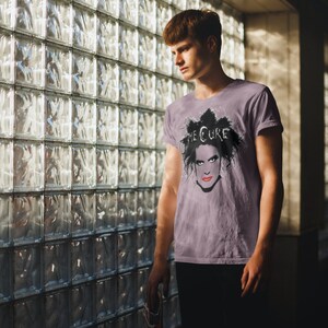 May include: A man wearing a purple t-shirt with a black and white image of Robert Smith, the lead singer of The Cure, on the front. The text "THE CURE" is printed above the image. The man is standing in front of a glass block wall.