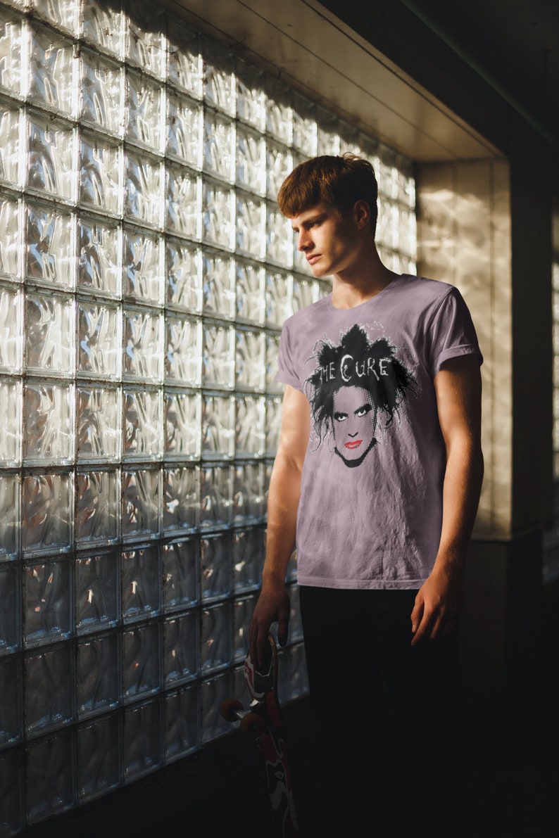 May include: A man wearing a purple t-shirt with a black and white image of Robert Smith, the lead singer of The Cure, on the front. The text "THE CURE" is printed above the image. The man is standing in front of a glass block wall.