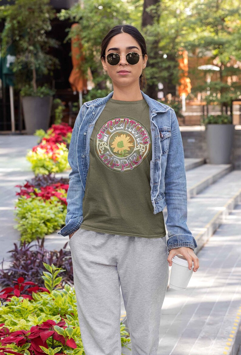 May include: A woman wearing a green t-shirt with a vintage graphic of a sunflower and the text "The Grateful Dead" in a circle. She is also wearing a denim jacket and grey sweatpants.