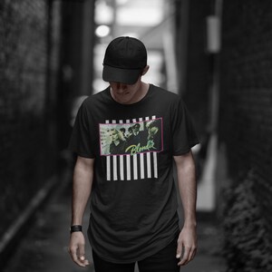 Op de afbeelding: Een persoon draagt een zwart T-shirt met een wit en zwart gestreept patroon en een afbeelding van een band genaamd "Blondie". De persoon draagt een zwarte baseballpet en staat in een smalle steeg.