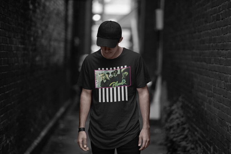 Op de afbeelding: Een persoon draagt een zwart T-shirt met een wit en zwart gestreept patroon en een afbeelding van een band genaamd "Blondie". De persoon draagt een zwarte baseballpet en staat in een smalle steeg.