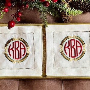 May include: Two white linen napkins with embroidered monograms in a circle. The monogram is "KBA" in red with a green and gold border. The napkins are on a gold tray.