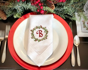 Puede incluir: Una servilleta de lino blanca con una corona bordada en rojo y verde y la letra "B" en el centro. La servilleta está doblada y colocada en un plato blanco con un borde rojo. El plato está sobre una mesa de madera marrón oscura.