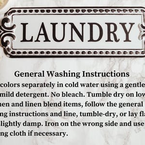May include: A white and black laundry sign with the word "LAUNDRY". The sign has decorative edges and includes general washing instructions. A wooden clothes peg is visible. The sign is on a marble surface.
