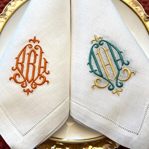May include: Two white linen napkins with embroidered monograms. One napkin features an orange monogram, the other a teal and gold monogram. The napkins are folded and placed on a white plate with a gold rim, ideal for a formal dinner setting.