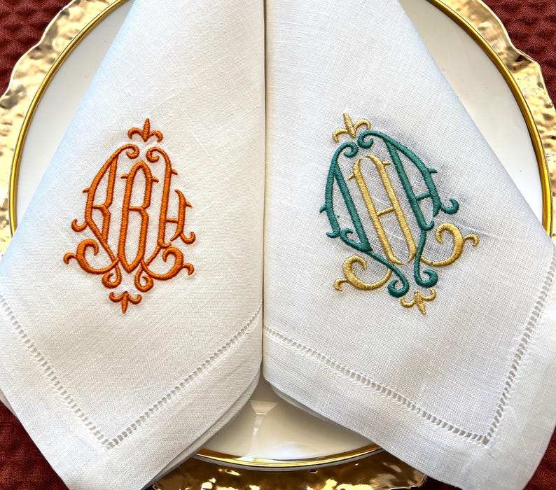 May include: Two white linen napkins with embroidered monograms. One napkin features an orange monogram, the other a teal and gold monogram. The napkins are folded and placed on a white plate with a gold rim, ideal for a formal dinner setting.