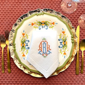 May include: A formal table setting with a white plate featuring a green and gold rim, floral design, and a monogrammed napkin. Gold cutlery and a gold-coloured spoon and knife are arranged on a textured red tablecloth.