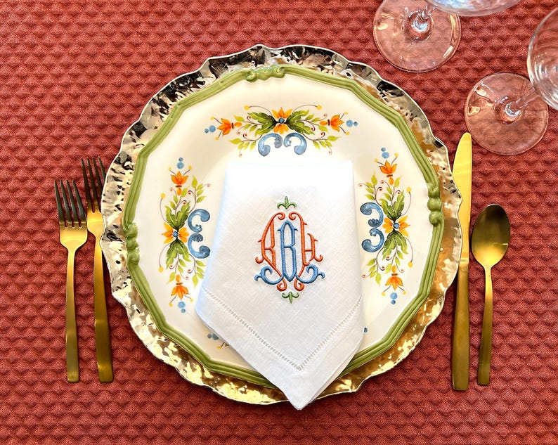 May include: A formal table setting with a white plate featuring a green and gold rim, floral design, and a monogrammed napkin. Gold cutlery and a gold-coloured spoon and knife are arranged on a textured red tablecloth.