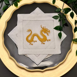 May include: White linen napkins with gold embroidered dragon designs. The napkins are folded and placed on a black and gold charger plate.