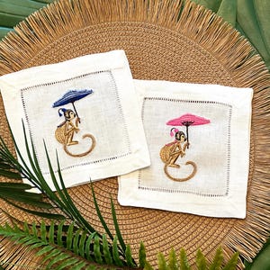 May include: Two white linen cocktail napkins with embroidered monkey designs. One monkey holds a blue umbrella, the other a pink umbrella. The napkins are placed on a woven straw mat with green foliage.