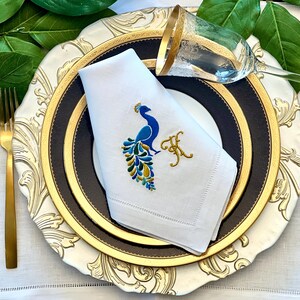 May include: Elegant table setting featuring a white napkin embroidered with a peacock design in blue and gold. The napkin rests on a layered plate with gold and black accents, complemented by gold cutlery and a wine glass.
