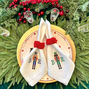 May include: A festive table setting featuring white linen napkins with embroidered nutcracker designs and red napkin rings. The napkins are placed on a decorative plate with a gold rim, surrounded by greenery and shot glasses, creating a holiday theme.