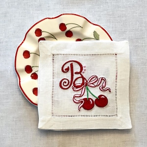 May include: A white linen cocktail napkin with a red embroidered design featuring the letter "B" and cherries. A cream-colored plate with a red cherry pattern is partially visible in the background.