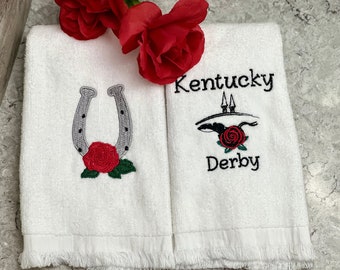 May include: Two white hand towels with embroidered designs. The first towel has a gray horseshoe with a red rose. The second towel has the text "Kentucky Derby" with a black silhouette of a horse and rider with a red rose.