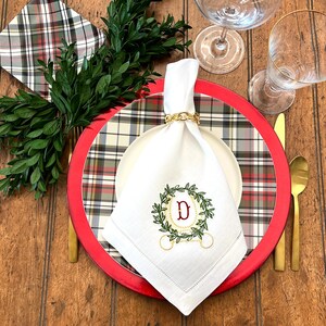 May include: A festive table setting with a white linen napkin featuring an embroidered wreath and the letter "D". The napkin is held by a gold chain napkin ring. The setting includes a red charger plate, plaid plate, and gold silverware.