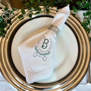 May include: White linen napkin with a green embroidered horseshoe and the letter "B" inside. The napkin is folded and placed on a silver charger.
