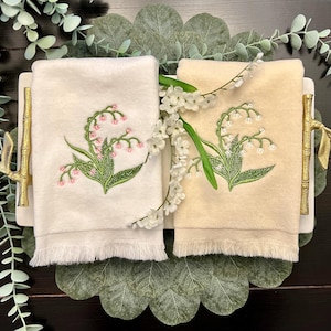 May include: Two folded hand towels, one cream and one off-white, with embroidered lily of the valley designs. The towels are displayed on a white tray with gold bamboo-style handles, surrounded by greenery and white flowers.