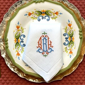 May include: A white linen napkin with an embroidered monogram in orange and blue, placed on a decorative plate. The plate features a floral design with green, orange, and blue accents, and a gold rim. Gold cutlery is visible.