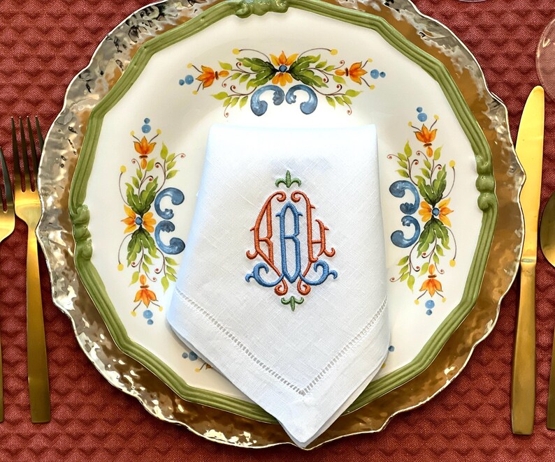 May include: A white linen napkin with an embroidered monogram in orange and blue, placed on a decorative plate. The plate features a floral design with green, orange, and blue accents, and a gold rim. Gold cutlery is visible.