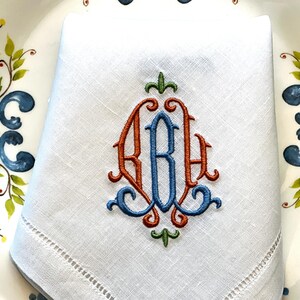 May include: White linen napkin with an embroidered monogram in orange, blue, and green. The napkin is folded and placed on a decorative plate with floral and leaf designs in blue, orange, and green.