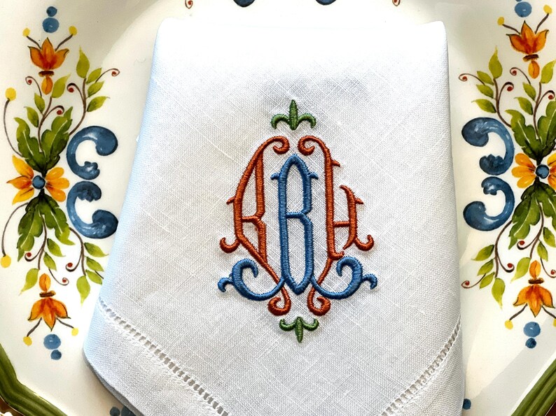 May include: White linen napkin with an embroidered monogram in orange, blue, and green. The napkin is folded and placed on a decorative plate with floral and leaf designs in blue, orange, and green.