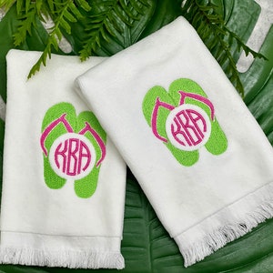 May include: Two white hand towels with fringed edges. Each towel features a green embroidered flip-flop with a pink monogram circle that reads "KRA".
