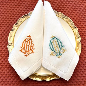 May include: Two white linen napkins, folded and placed on a gold-rimmed plate. One napkin has an orange monogram, the other a teal and gold monogram. The napkins are on a white plate with a gold rim.