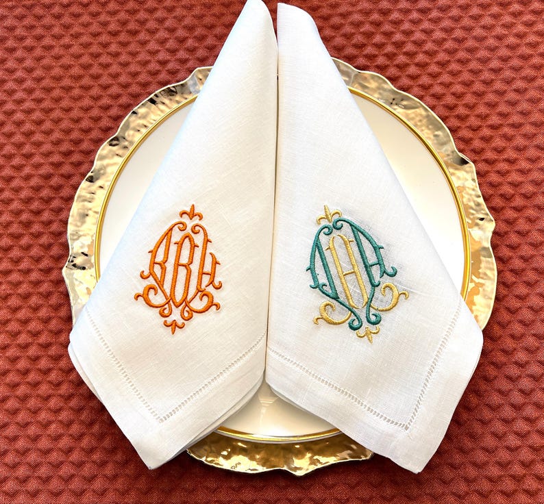 May include: Two white linen napkins, folded and placed on a gold-rimmed plate. One napkin has an orange monogram, the other a teal and gold monogram. The napkins are on a white plate with a gold rim.