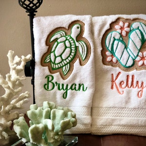 May include: Two white towels with embroidered designs. The towel on the left has a green sea turtle with a brown outline and the name "Bryan" embroidered in green. The towel on the right has a pair of green flip flops with brown outlines and pink flowers and the name "Kelly" embroidered in orange.