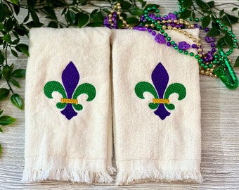 Könnte beinhalten: Zwei weiße Handtücher mit einem gestickten lila und grünen Fleur-de-lis auf jedem. Die Handtücher haben einen Fransenrand.