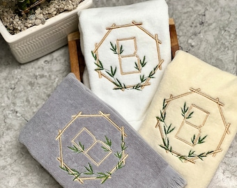 May include: Three towels with embroidered monograms in a gold frame with green leaves. The towels are in shades of gray, white, and cream.