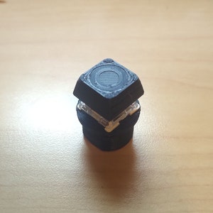 May include: A black bottle stopper with a geometric design. The stopper has a square top with a circular pattern and a cylindrical base. The stopper is made of a dark material and has a clear, plastic-like section.