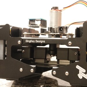 May include: A black metal device with the text "DingKey Designs" and a logo. The device has several electronic components, including a circuit board, motor, and wiring. The device is mounted on a black frame with silver screws.