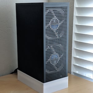 May include: A black and gray computer case with a perforated front panel and two visible blue fans. The case is rectangular and sits on a white base. The case is on a light wood surface.